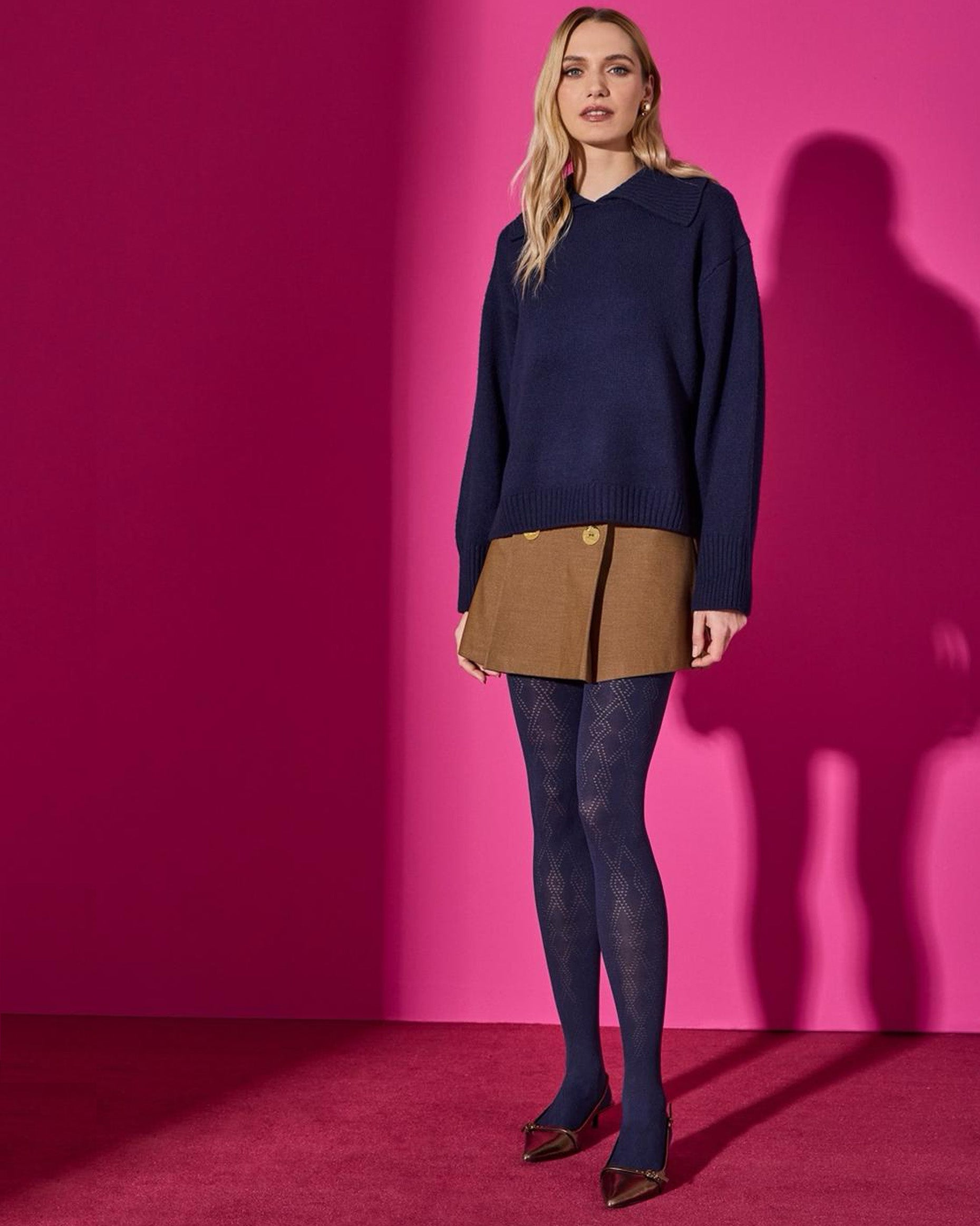 Woman wearing a navy sweater, brown skirt and diamond patterned tights against a pink wall