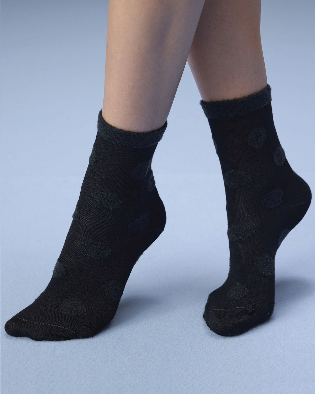 Black ankle socks with a subtle tonal polka dot pattern, shown on a model's feet against a light blue studio background.