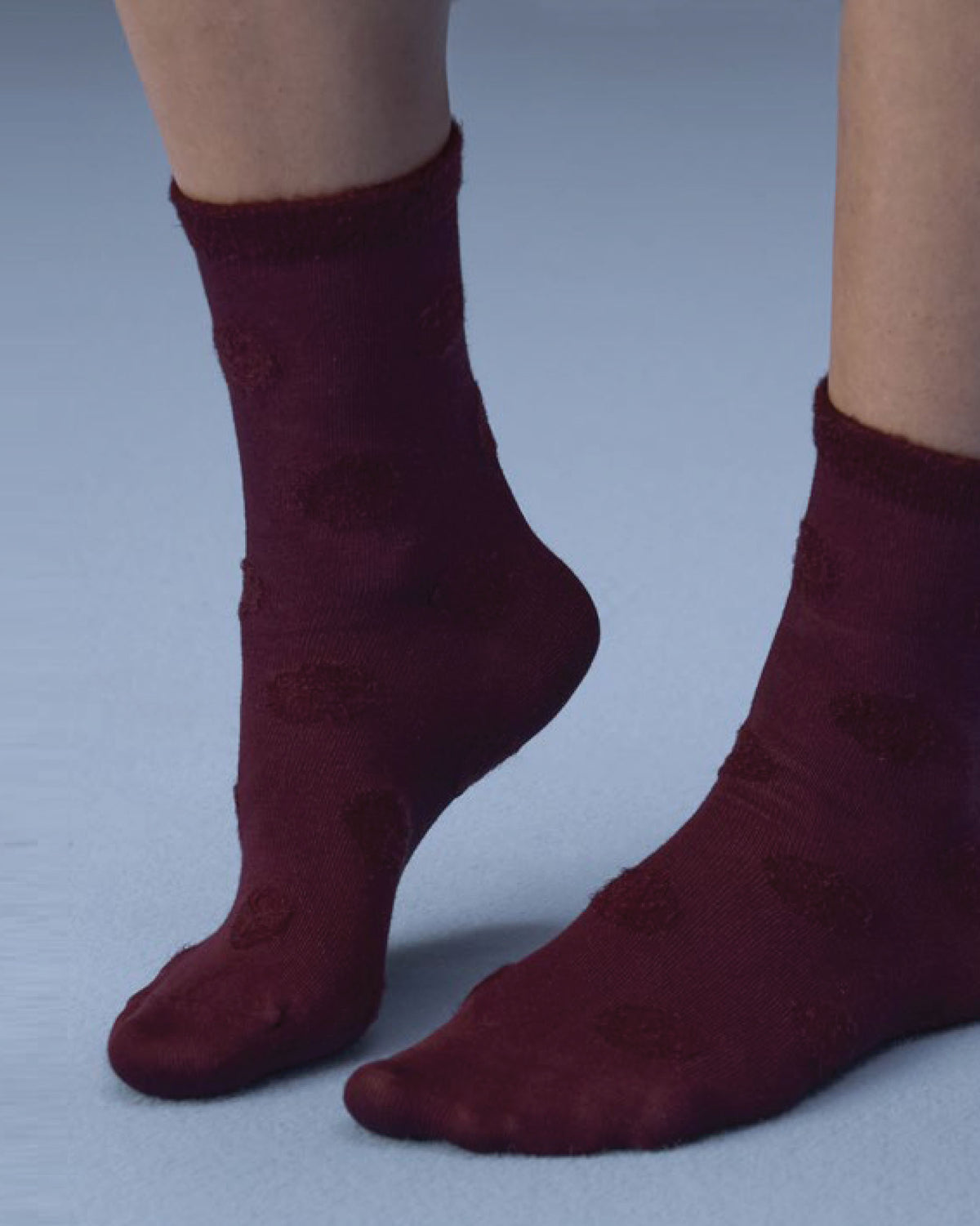 A close-up of a person's feet wearing dark burgundy socks with a subtle tonal textured pattern, set against a solid light blue-gray background.