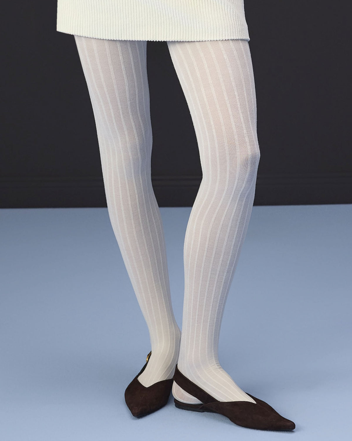 White ribbed tights worn by a model, paired with dark brown pointed flats, shown from the waist down against a dark gray and light blue studio background.