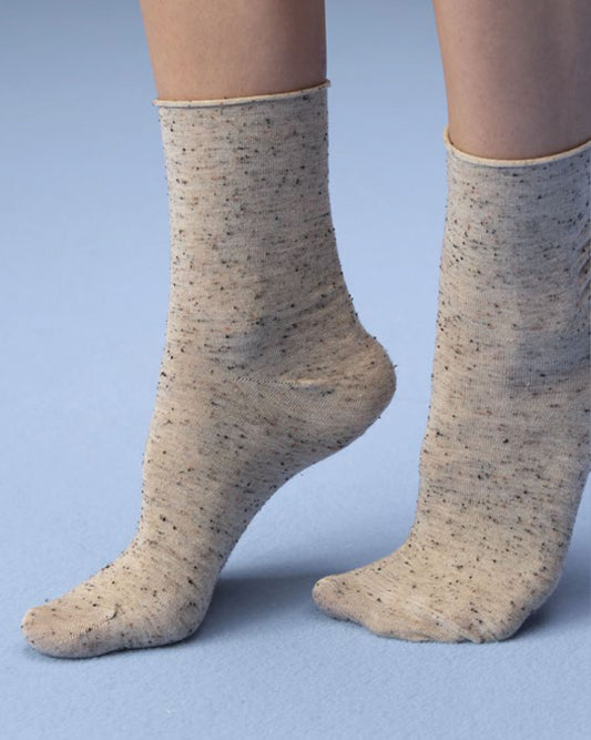 A pair of beige knit socks with small black flecks, worn on a person's feet against a light blue background.