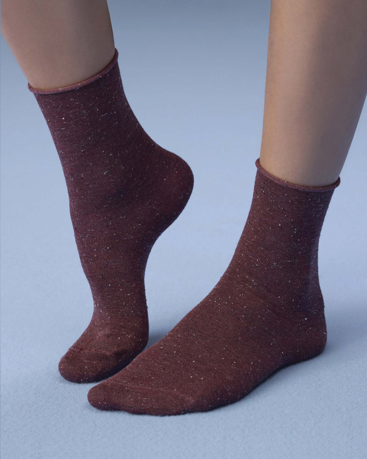 A pair of ankle-high socks in a dark reddish-brown color with subtle white fleck, shown on a model's feet against a light blue-gray background.