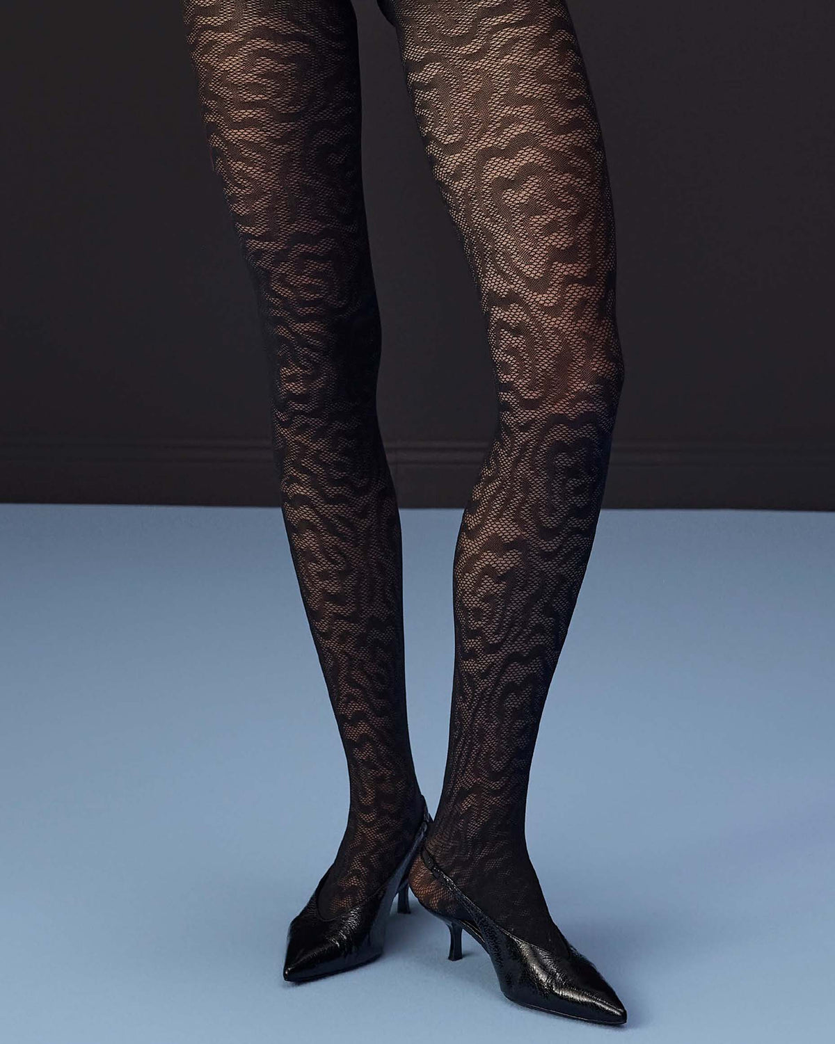 Black patterned tights worn on legs with black pointed-toe heels, photographed against a dark gray and light blue studio background.