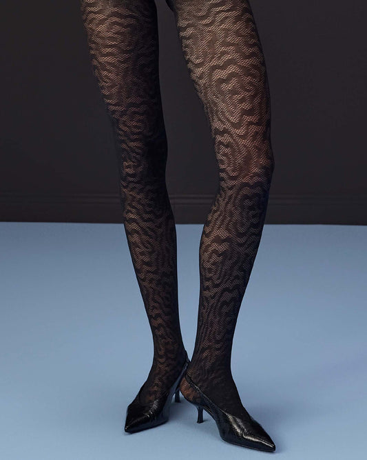 Black patterned tights worn on legs with black pointed-toe heels, photographed against a dark gray and light blue studio background.