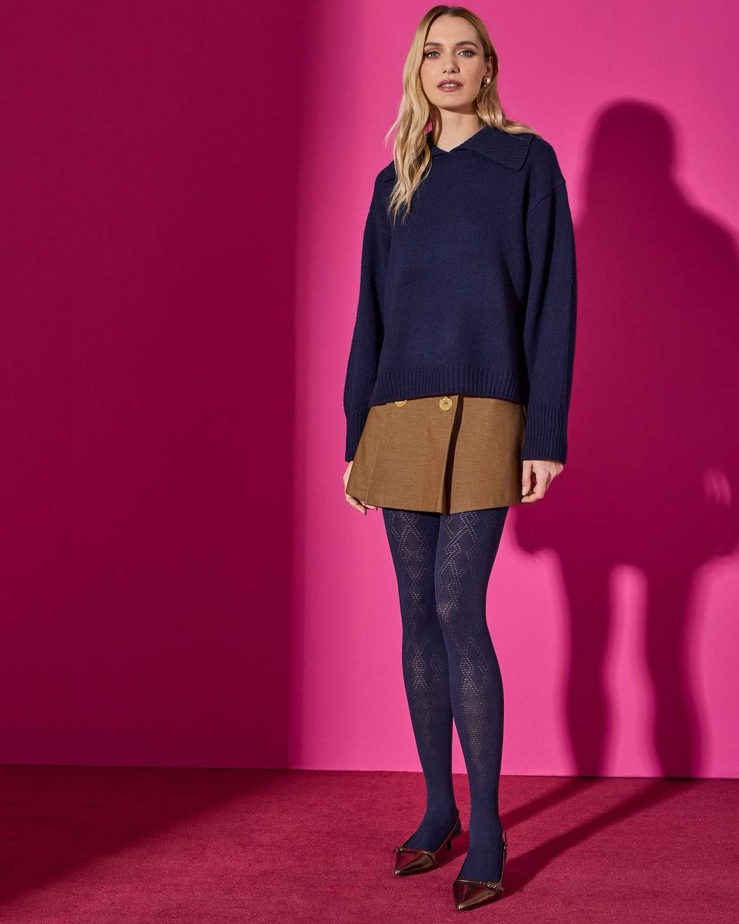 Woman wearing a navy sweater, brown skirt and diamond patterned tights against a pink wall