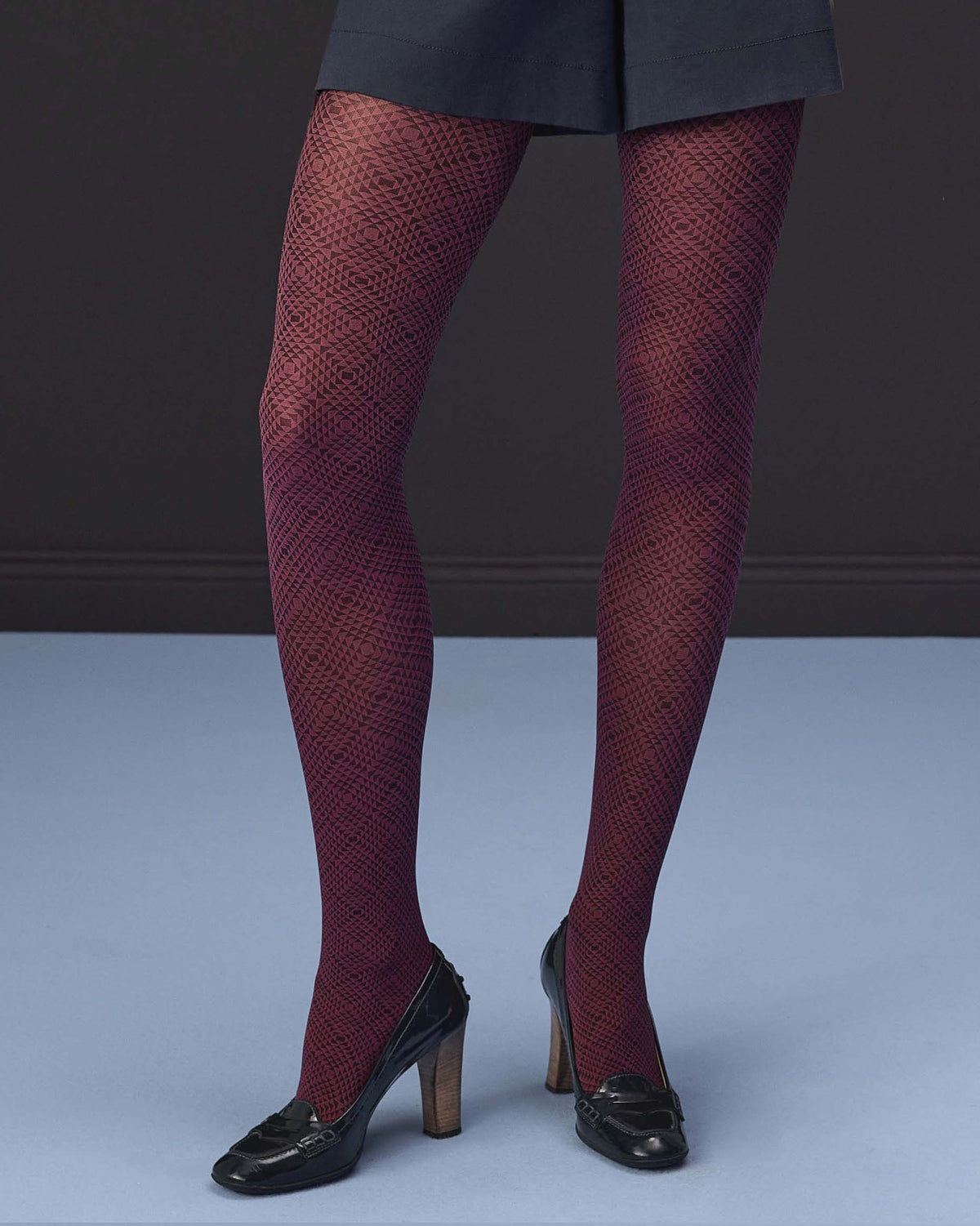 A person from the waist down wearing dark burgundy patterned tights with a black skirt and black high heels, standing against a dark gray wall and light blue floor.