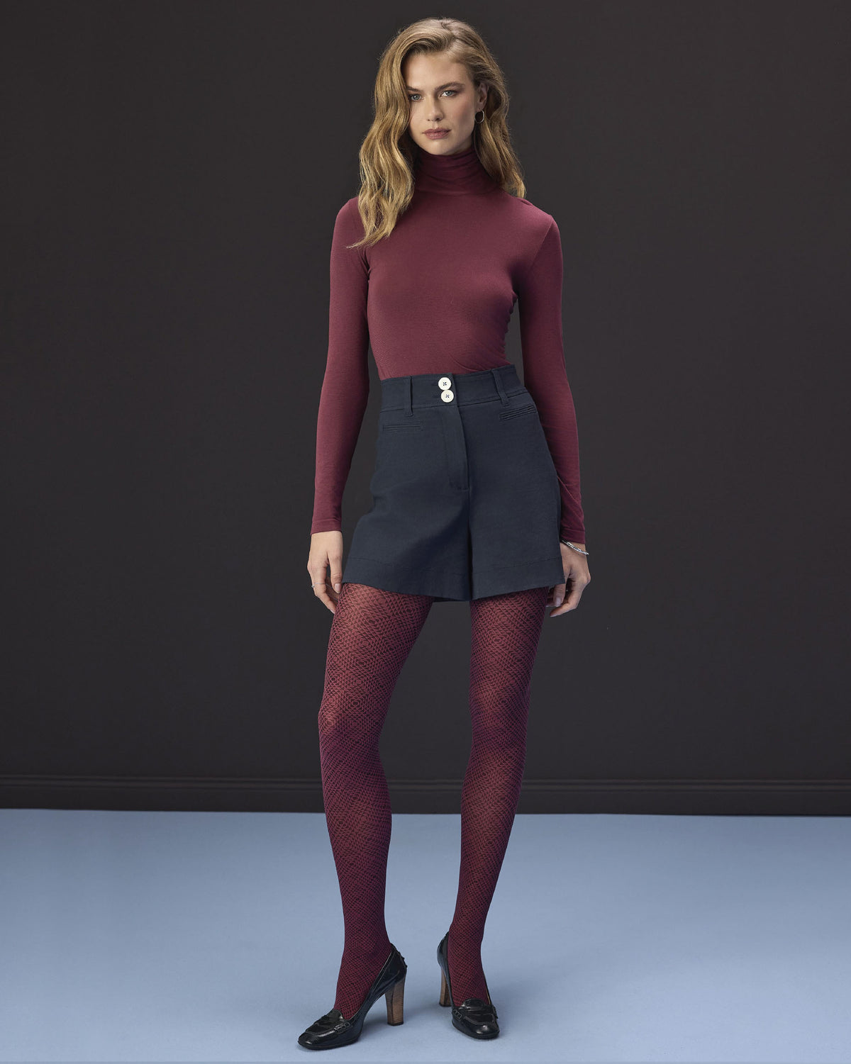 A model stands against a dark gray studio background wearing a fitted burgundy mock-neck long-sleeve top, a dark slate blue mini skirt with a silver button, matching burgundy patterned tights, and black heels.