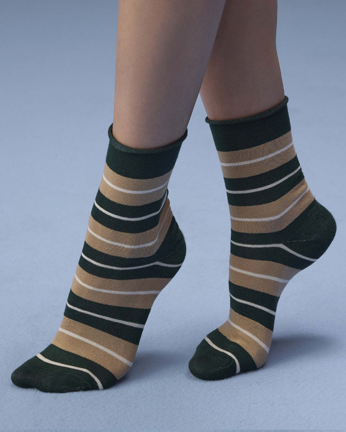 A pair of crew socks with horizontal stripes in dark green, tan, and white, worn on a person's feet against a light blue-gray studio background.